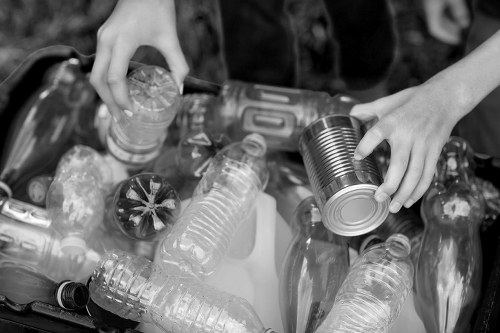 A pair of hands are sorting through a collection of various waste items, primarily plastic bottles and containers, on top of a surface. The plastic bottles include transparent and dark-colored ones, some with visible labels or caps, and are arranged in a somewhat scattered manner. One hand is holding a cylindrical metal can, while the other hand is grasping a plastic bottle by its neck. The background shows additional waste materials, and the overall scene emphasizes waste sorting and disposal, typical of rubbish removal activities. The image captures the textures of plastic and metal surfaces and highlights the contrast between different types of containers, aligning with the context of professional rubbish collection in the local Redbridge area.