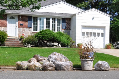 A residential house with a white garage door and brick exterior walls, situated in a suburban area. In the foreground, several large grey and beige rocks are lined up along the curb, with a white plastic container holding dried and dead plant stems placed among them. The house has a front lawn with well-maintained green grass, small bushes, and a paved pathway leading to the front door, which is located on the left side of the house. The background features a blue sky with some trees visible behind the house. This scene reflects a typical suburban environment where rubbish clearance or garden waste removal services by Commercial Waste Redbridge might be required to handle landscape debris and curbside waste in the local area near Redbridge.