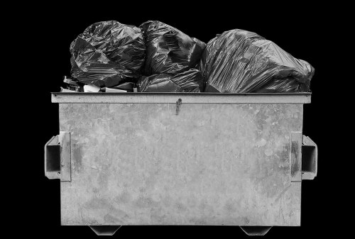 A large horizontally oriented industrial skip filled with black garbage bags, primarily made of plastic, with a textured, crinkled appearance. The bags are stacked towards the top of the skip, some leaning to the left and others to the right, filling the container almost to the rim. The skip, constructed of sturdy, grey, weathered steel with visible scratches and slight discoloration, is positioned outdoors on a flat surface, likely a driveway or pavement in a commercial or residential area within Redbridge. The background is black, emphasizing the container and its contents without any additional environmental context. The image, associated with rubbish removal services by Commercial Waste Redbridge, illustrates the typical type of waste collected and disposed of by local rubbish clearance providers, highlighting the capacity and utilitarian design of commercial waste containers used for significant rubbish clearance tasks.