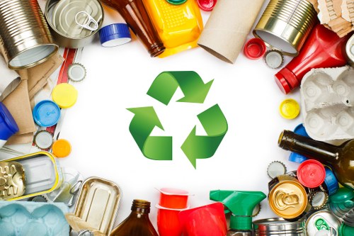 A diverse collection of household waste items arranged in a circular pattern around a large green recycling symbol. Visible objects include empty aluminum cans with smooth, reflective metallic surfaces; plastic bottles in various colors such as red, green, yellow, and brown with textured labels and caps; cardboard tubes with corrugated surfaces and brown coloring; crumpled white paper or paperboard waste; plastic containers and jugs of different sizes and colors, some with handles; and small plastic caps and lids scattered throughout. The background is white, emphasizing the waste materials and the central recycling icon, representing the importance of rubbish and waste management services in Redbridge. The arrangement highlights different materials for recycling, within the context of rubbish removal for commercial or domestic purposes. The environment appears clean and organized, suitable for illustrating waste disposal and recycling efforts in the local area served by Commercial Waste Redbridge.