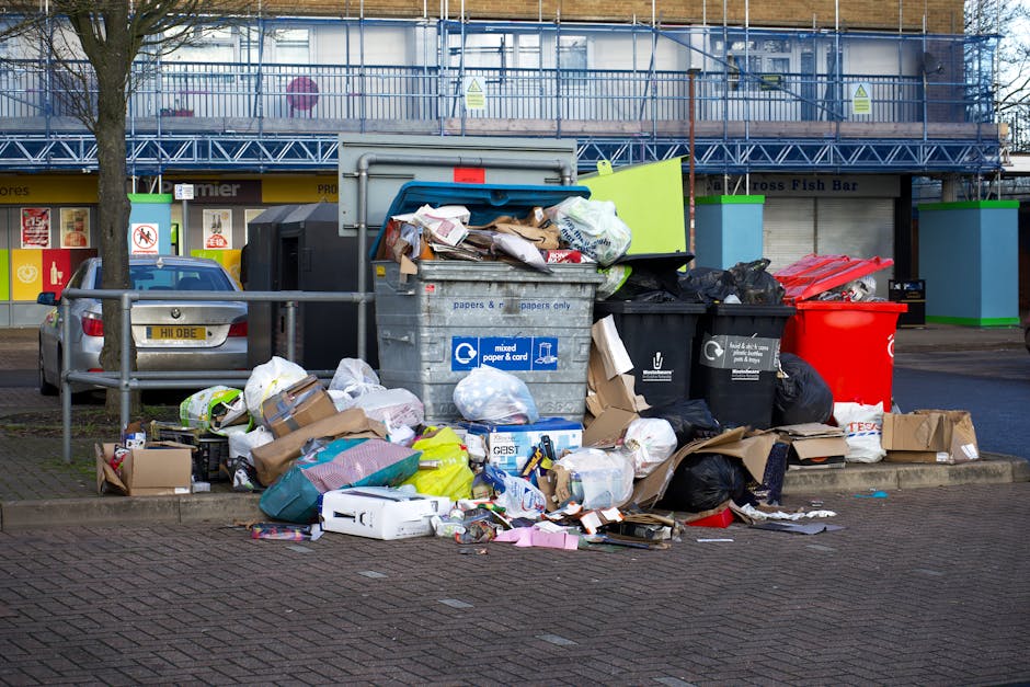 A large outdoor collection point for domestic and commercial waste situated on a paved urban street. In the foreground, there is an overflowing grey recycling bin marked for mixed paper and cardboard,