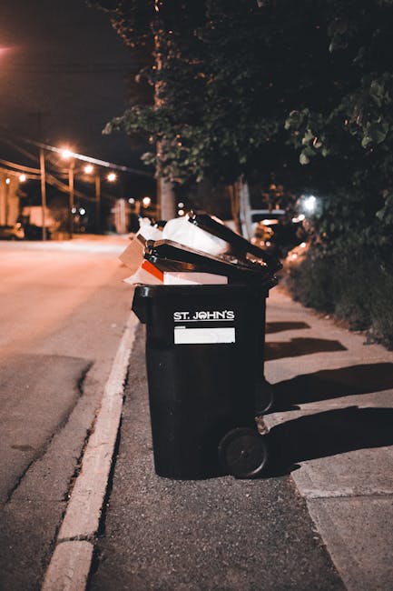 A large outdoor collection point for domestic and commercial waste situated on a paved urban street. In the foreground, there is an overflowing grey recycling bin marked for mixed paper and cardboard,